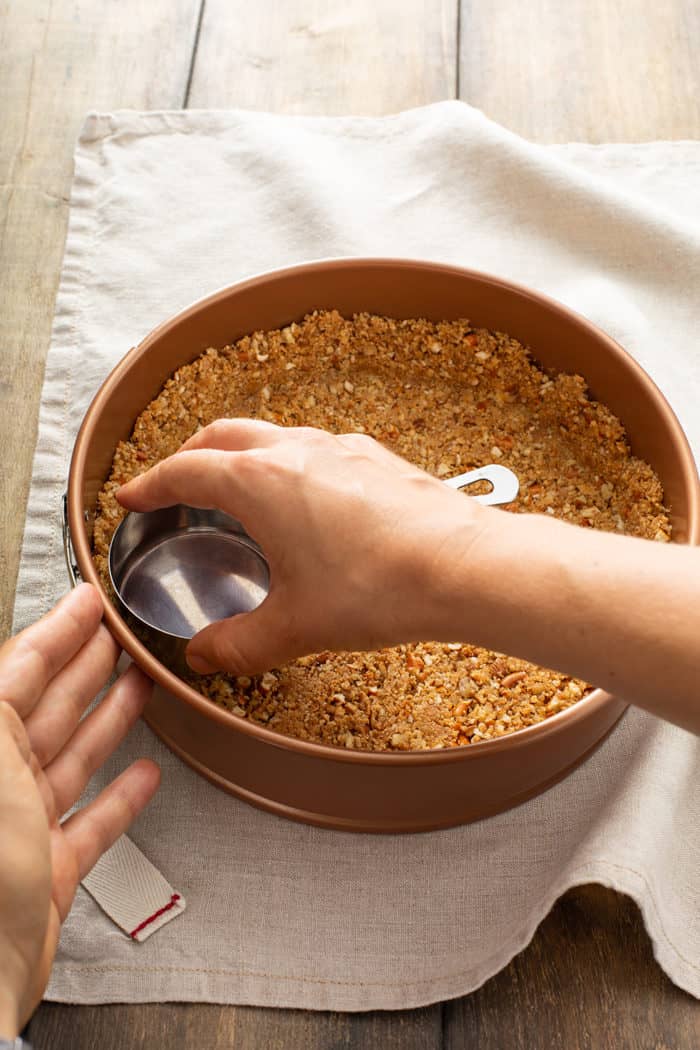 Hand using a metal measuring cup to press a pretzel and graham cracker pie crust into a springform pan