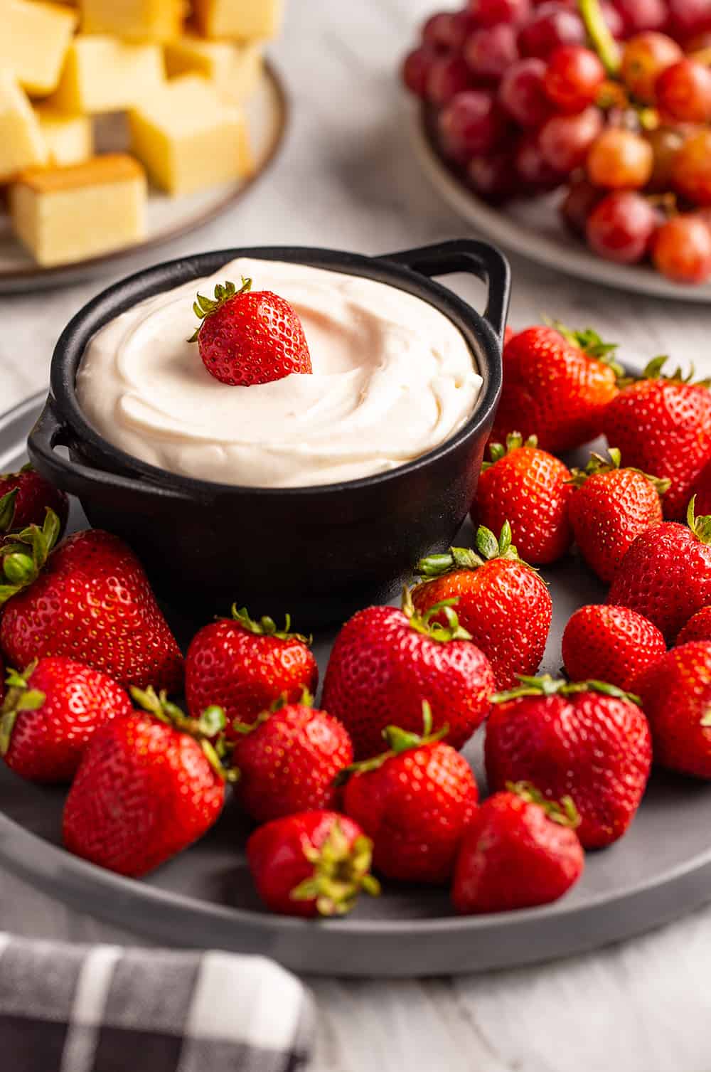 Strawberries surrounding a bowl of fruit dip on a black plate. A whole strawberry sits on top of the dip.