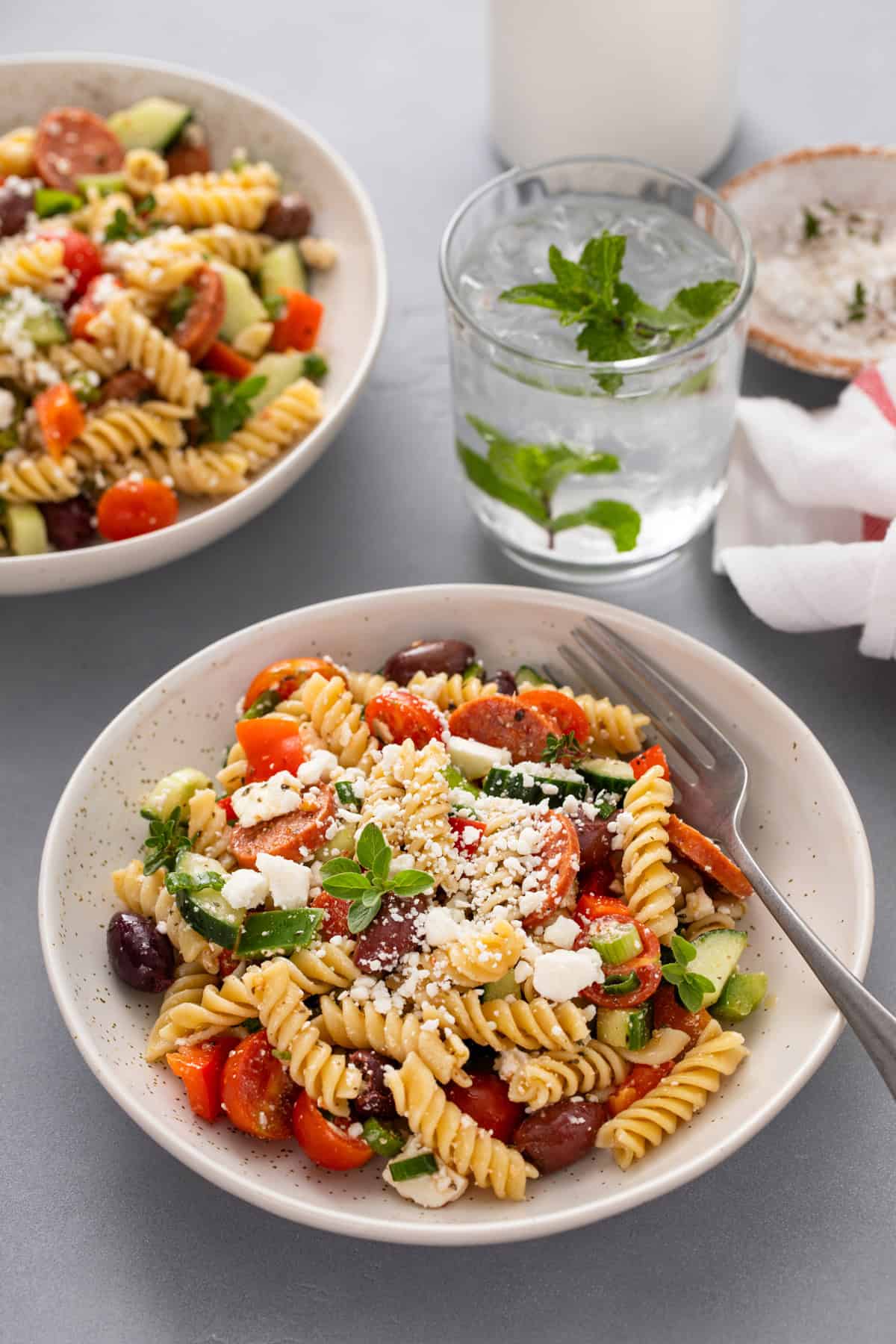 Two bowls filled with greek pasta salad with a glass of water in the background.