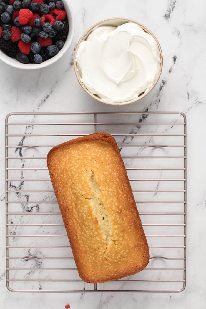 Overhead view of lemon whipping cream cake cooling on a wire rack next to a bowl of whipped cream and fresh berries