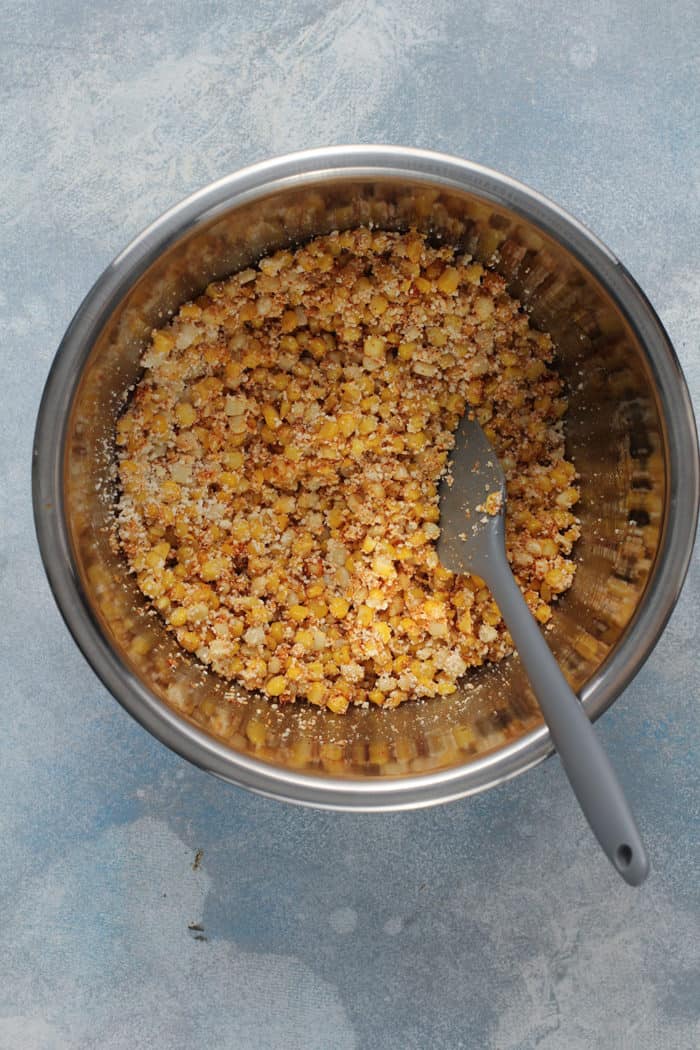 Spatula combining charred corn, cotija cheese and chili powder in a metal bowl