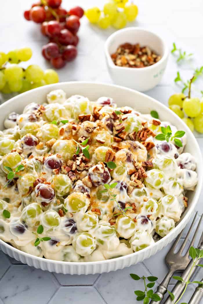 3/4 view of creamy grape salad in a white serving bowl, surrounded by grapes and a bowl of pecans