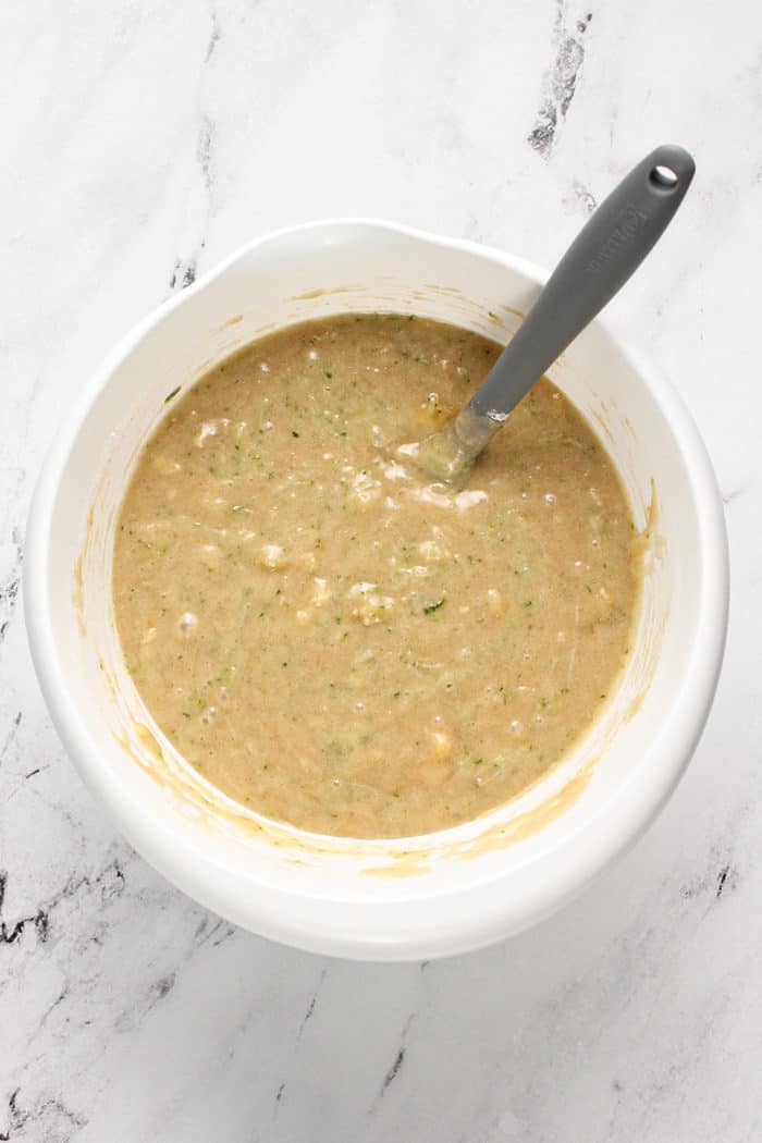 Zucchini banana bread batter being stirred by a spatula in a white mixing bowl