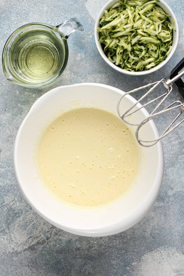 Mixer beating together eggs and sugar and about to add oil and zucchini in a white mixing bowl