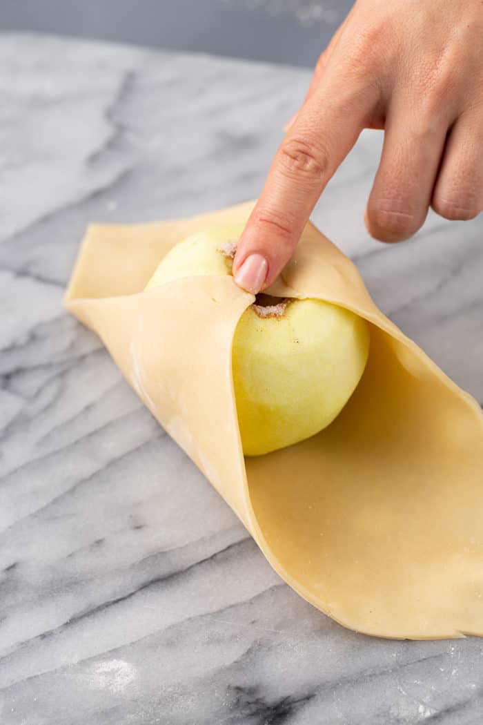 Hand bringing the corners of a square of pie dough up and over a peeled and cored apple to form an apple dumpling