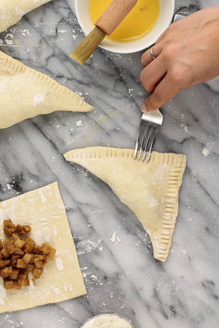 Fork crimping the edges of an assembled apple turnover on a marble countertop