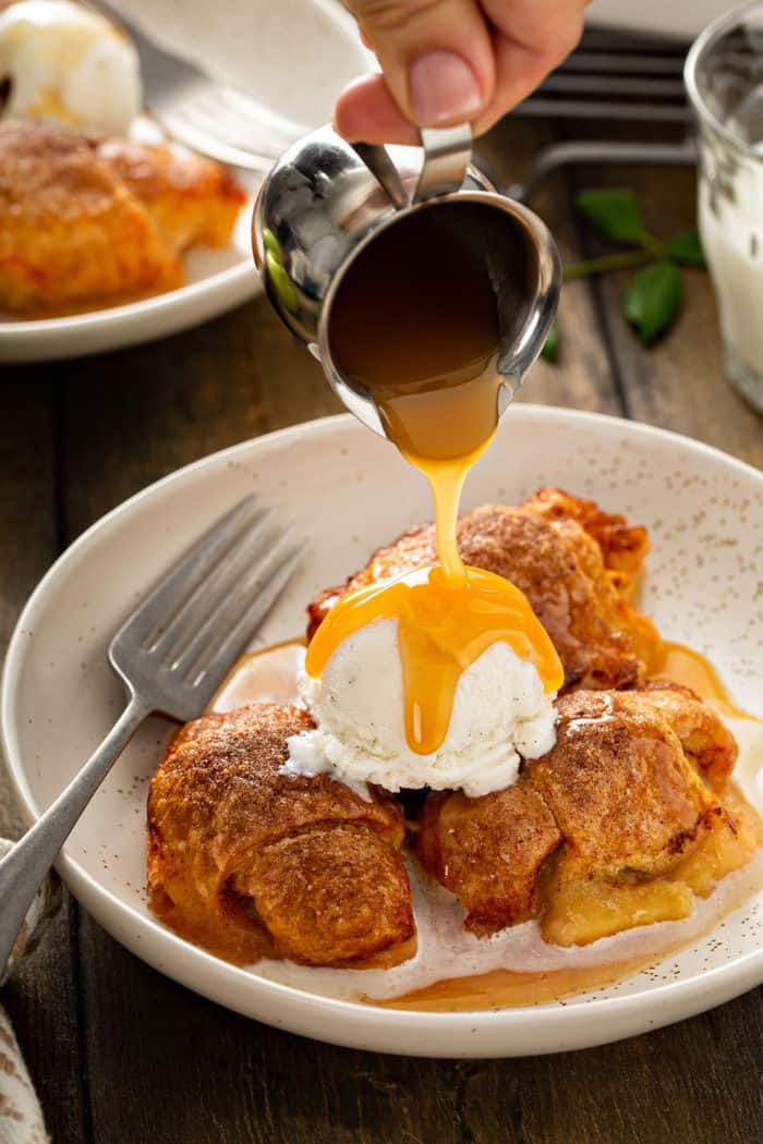 Caramel sauce being poured over easy apple dumplings and a scoop of vanilla ice cream in a cream bowl