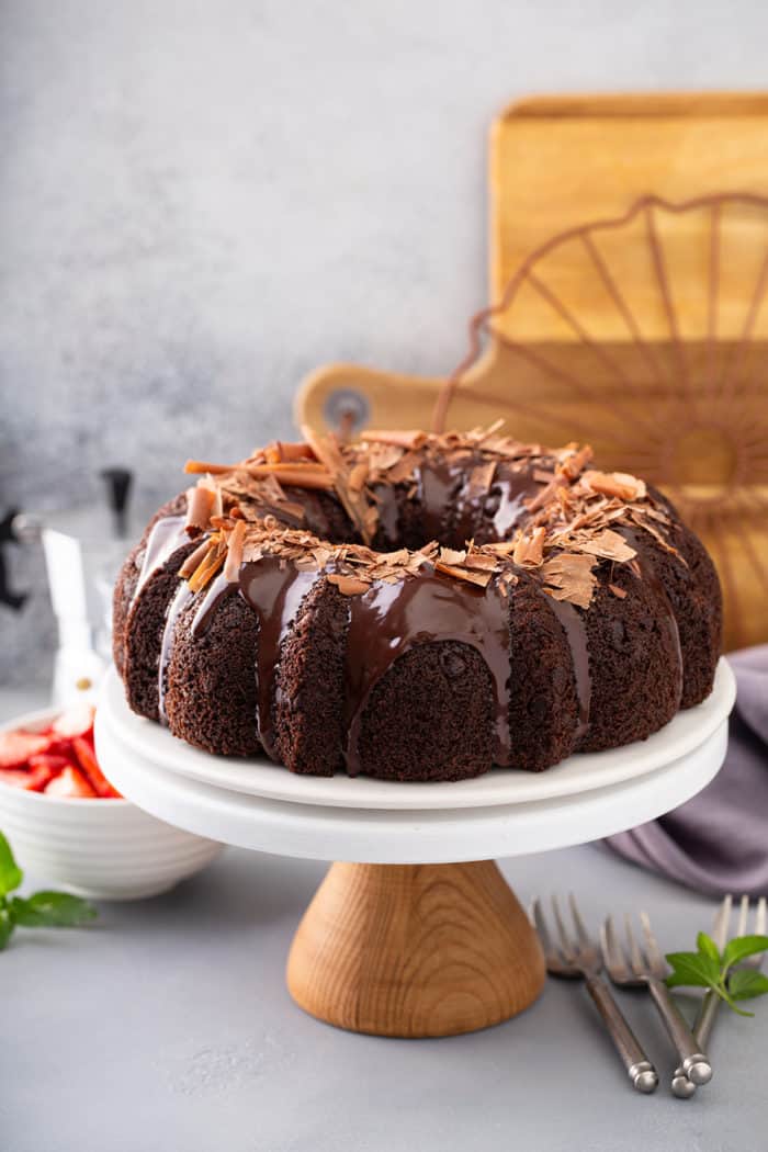 Chocolate bundt cake topped with ganache and chocolate shavings on a cake plate with berries and espresso in the background