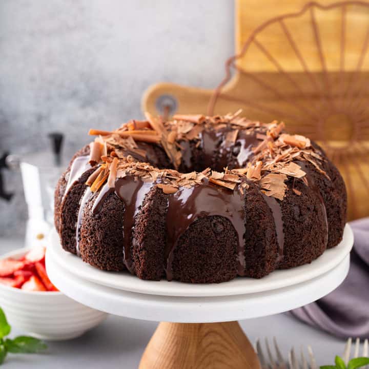 Chocolate bundt cake topped with ganache and chocolate shavings on a cake plate with berries and espresso in the background