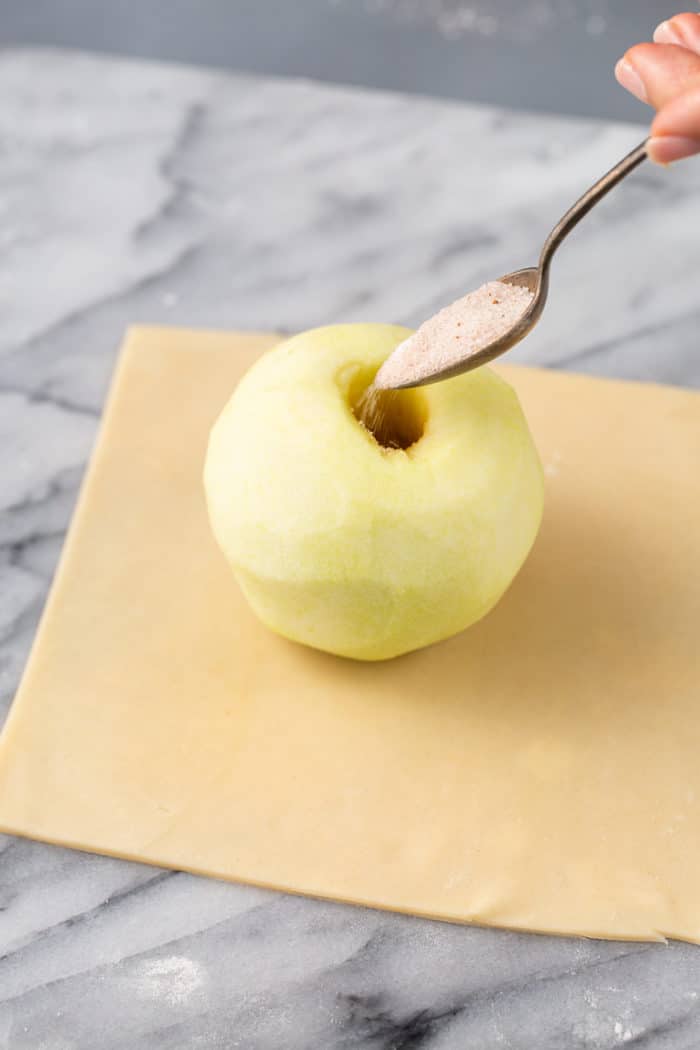 Spoon pouring spiced sugar into the center of a peeled and cored apple that is set in the center of a square of pie dough