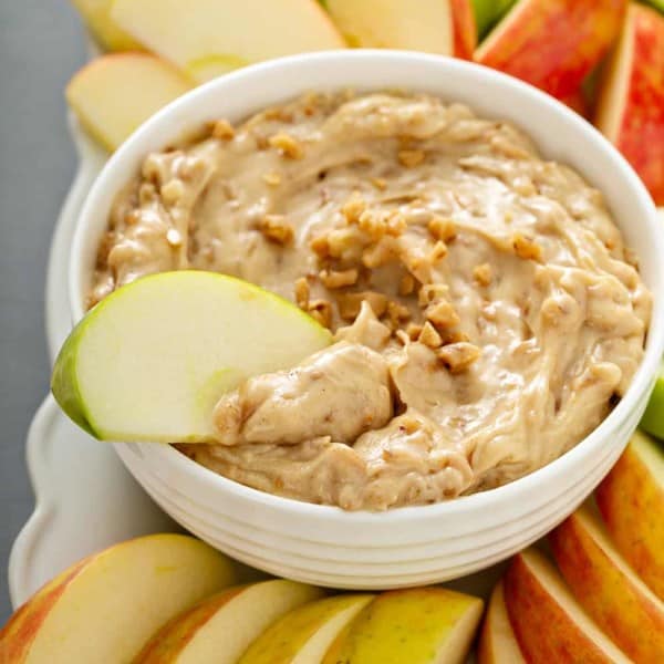 Apple slice dipped into a white bowl of toffee apple dip, surrounded by sliced apples