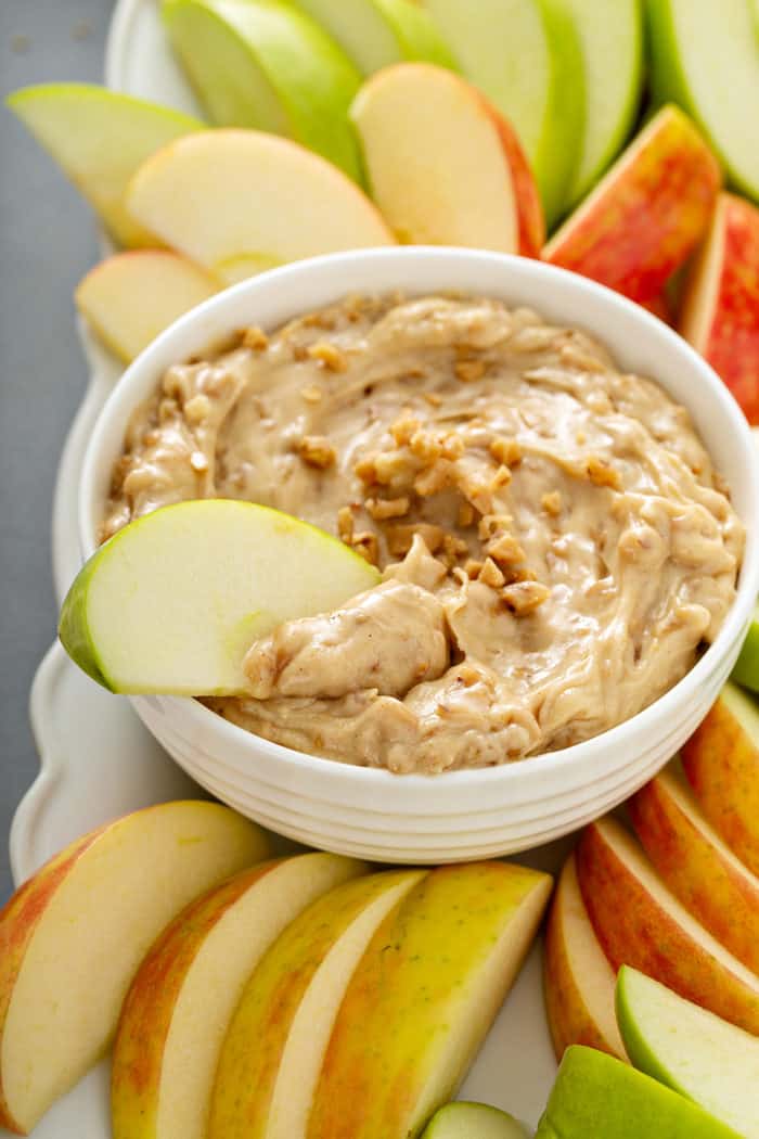 Apple slice dipped into a white bowl of toffee apple dip, surrounded by sliced apples