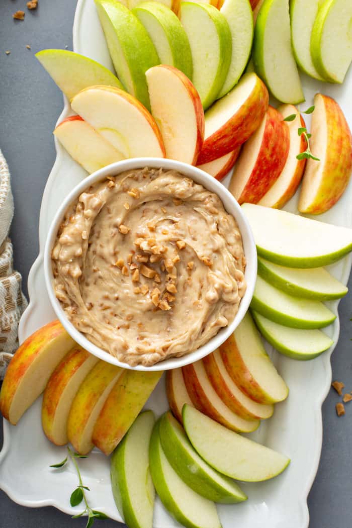 Overhead view of a bowl of toffee apple dip surrounded by fresh apples on a white platter