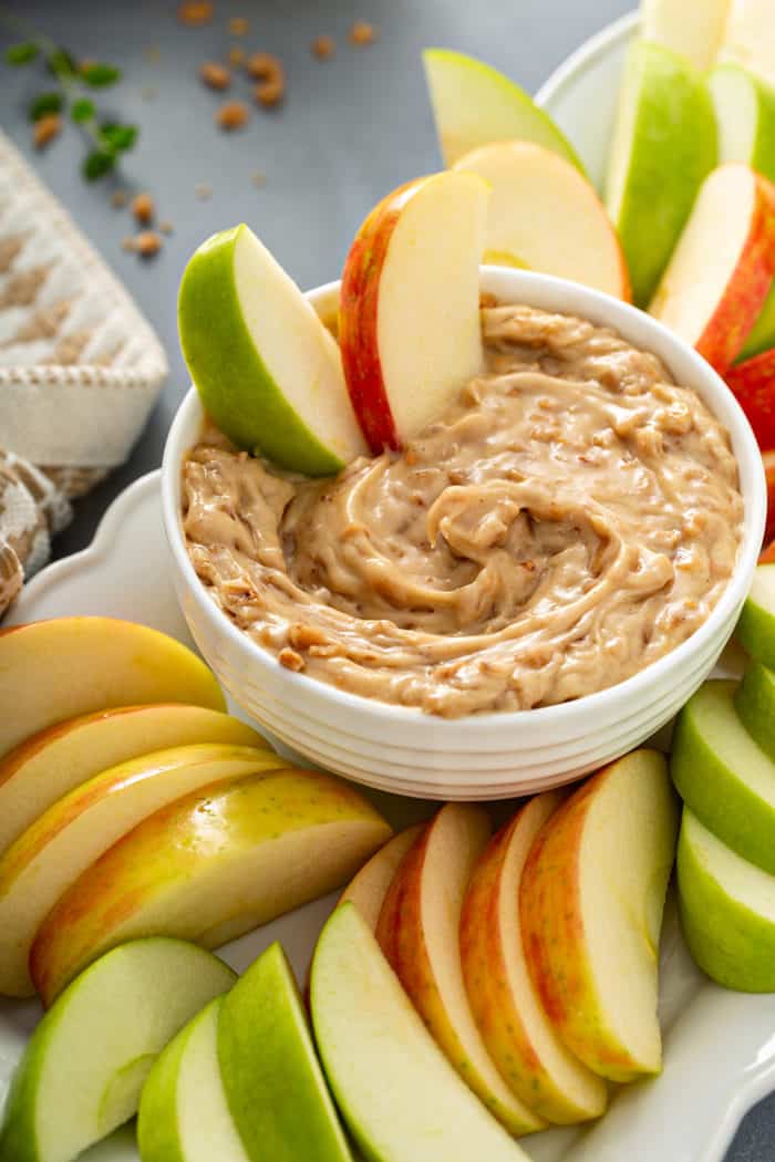 Slices of fresh apples in a bowl of toffee apple dip, surrounded by sliced apples on a platter