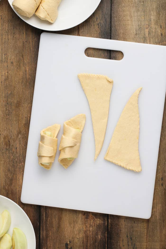 Apple slices being wrapped in triangles of crescent roll dough on a white cutting board