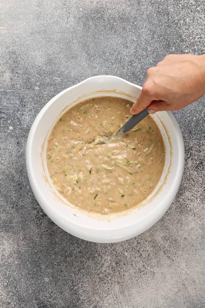 Hand stirring zucchini cake batter in a white mixing bowl with a gray spatula