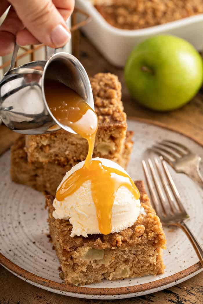 Caramel sauce being poured over a sliced of apple coffee cake topped with ice cream