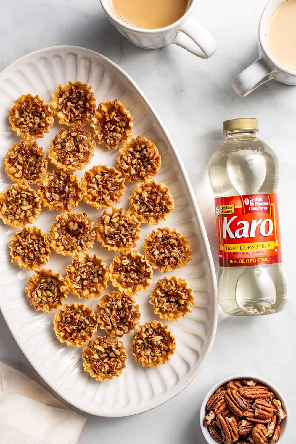 Overhead view of mini pecan pies on a white platter, set next to a bottle of corn syrup on a countertop