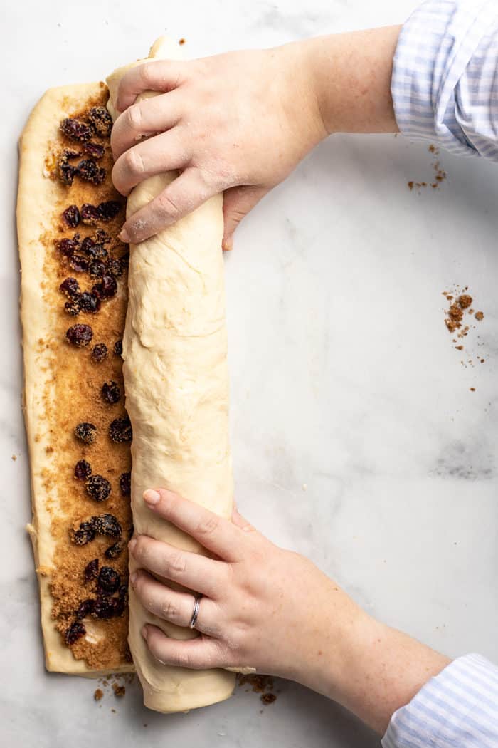 Hands rolling cranberry orange cinnamon roll dough into a log on a marble countertop