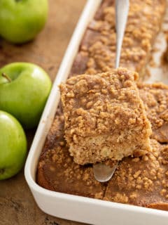 Cake server holding a slice of apple coffee cake over a pan of coffee cake