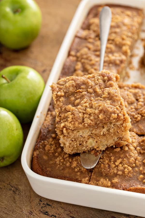 Cake server holding a slice of apple coffee cake over a pan of coffee cake