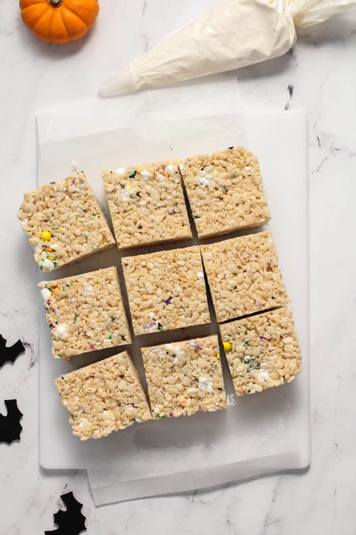 Unfrosted halloween rice krispie treats, sliced and set on a piece of parchment next to a bag of buttercream frosting