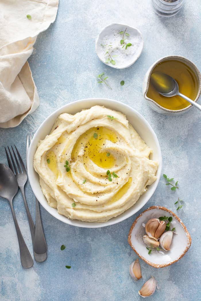 Bowl of garlic mashed potatoes on a blue countertop next to garlic cloves and melted butter