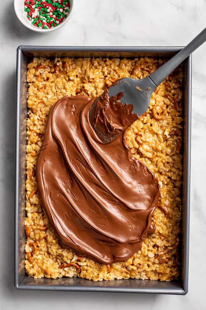 Spatula spreading chocolate on top of peanut butter pretzel bars in a metal pan