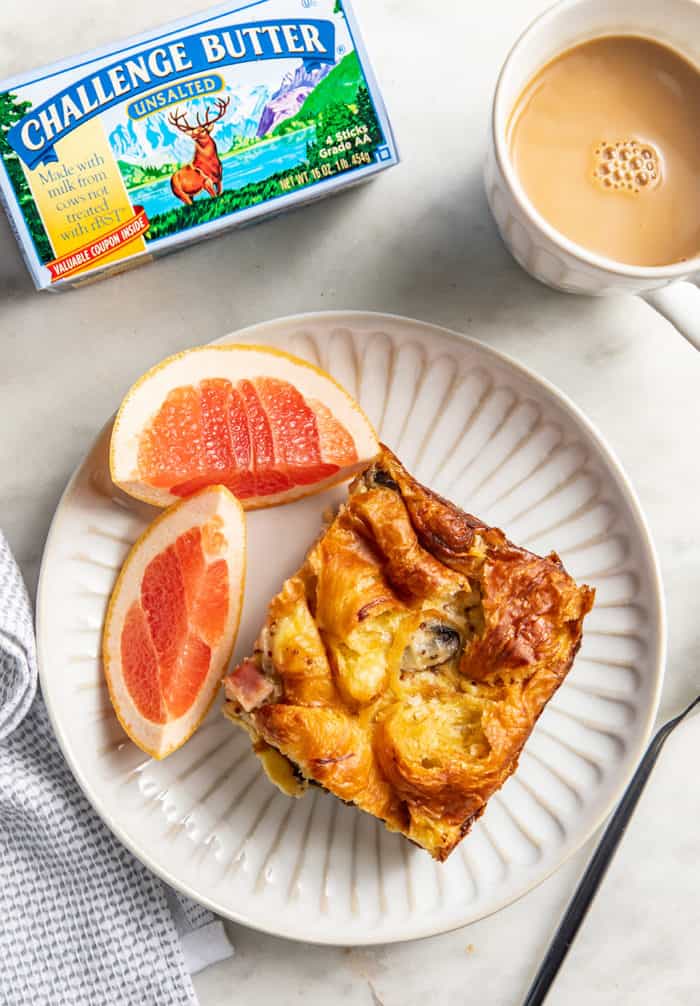 Overhead view of a slice of croissant breakfast casserole on a plate alongside two pieces of grapefruit