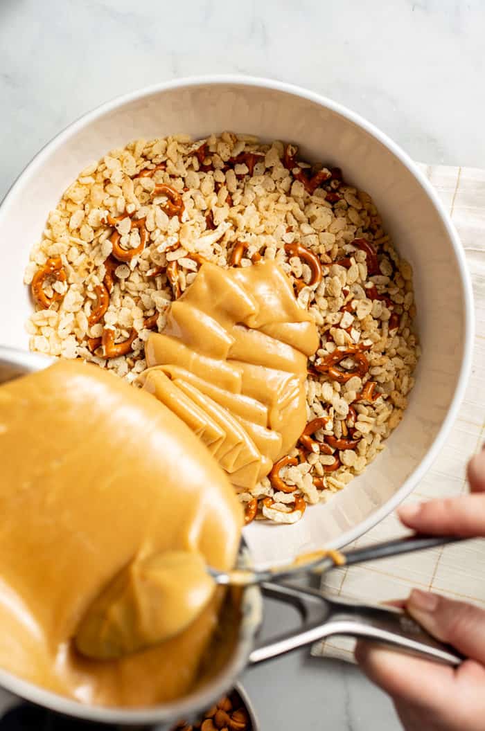 Peanut butter syrup being poured over rice cereal and pretzels in a white mixing bowl