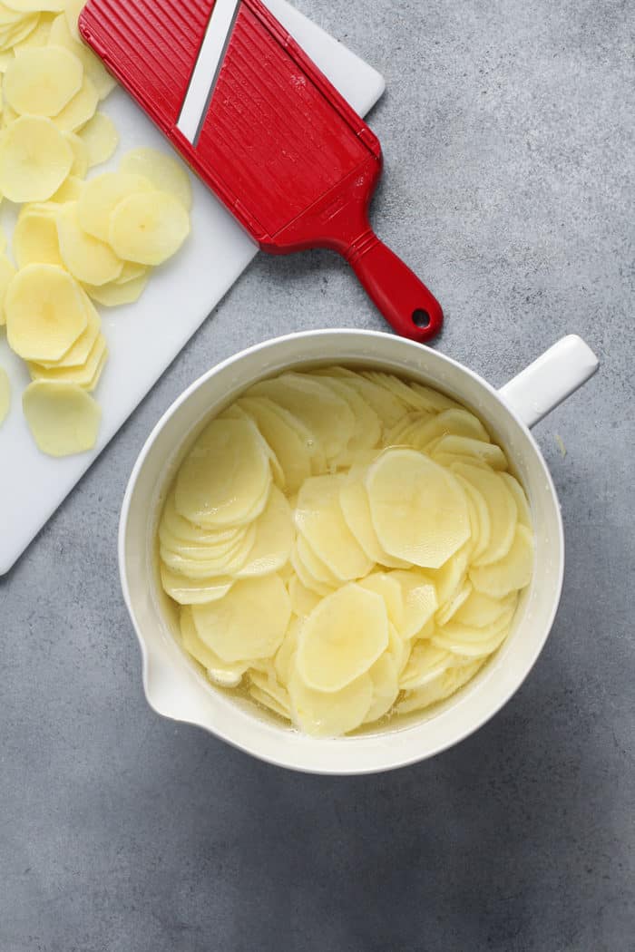 thinly sliced potatoes soaking in cold water in a white bowl