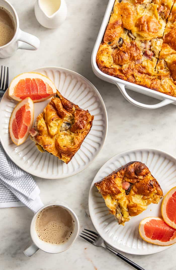 Two white plates holding slices of croissant breakfast casserole and slices of grapefruit alongside a baking dish of breakfast casserole