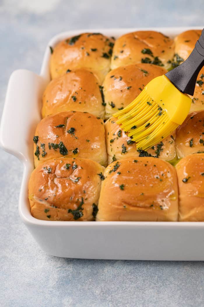 Butter and herbs being brushed over the top of hawaiian rolls for sliders in a white pan