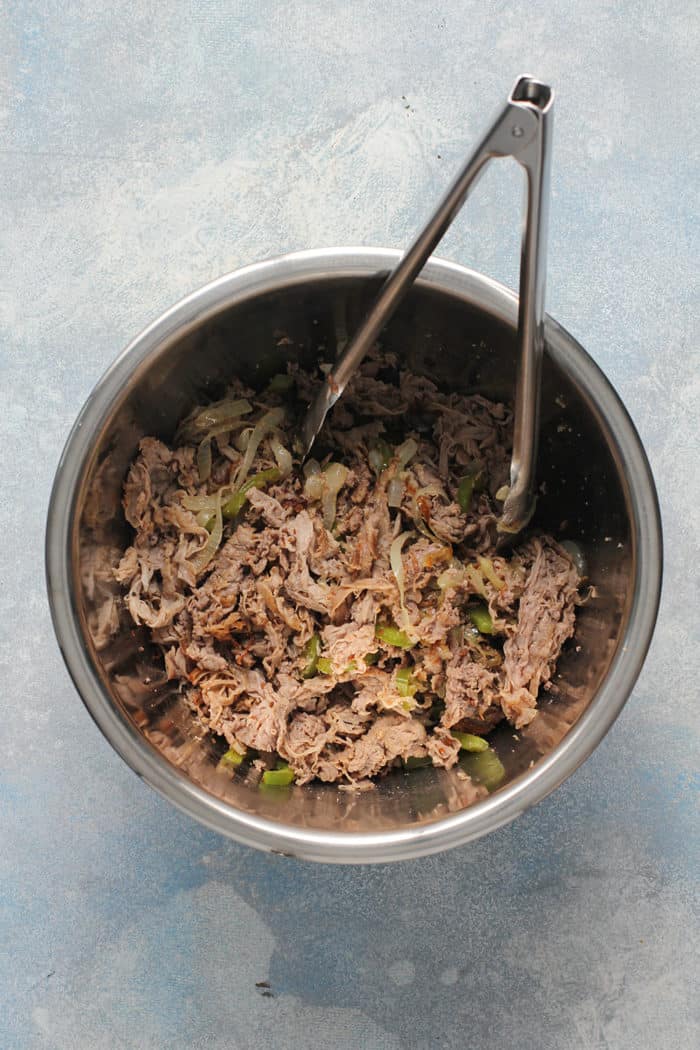 Tongs stirring cooked shaved beef, onions, and peppers in a metal bowl