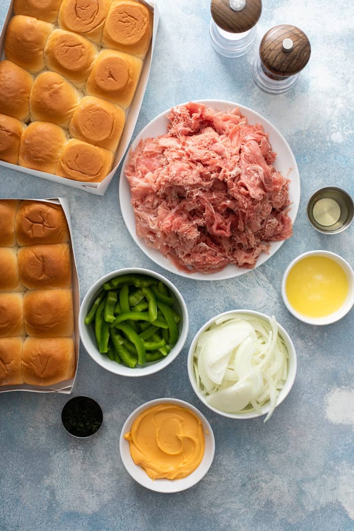 Overhead view of ingredients for cheesesteak sliders arranged on a blue countertop