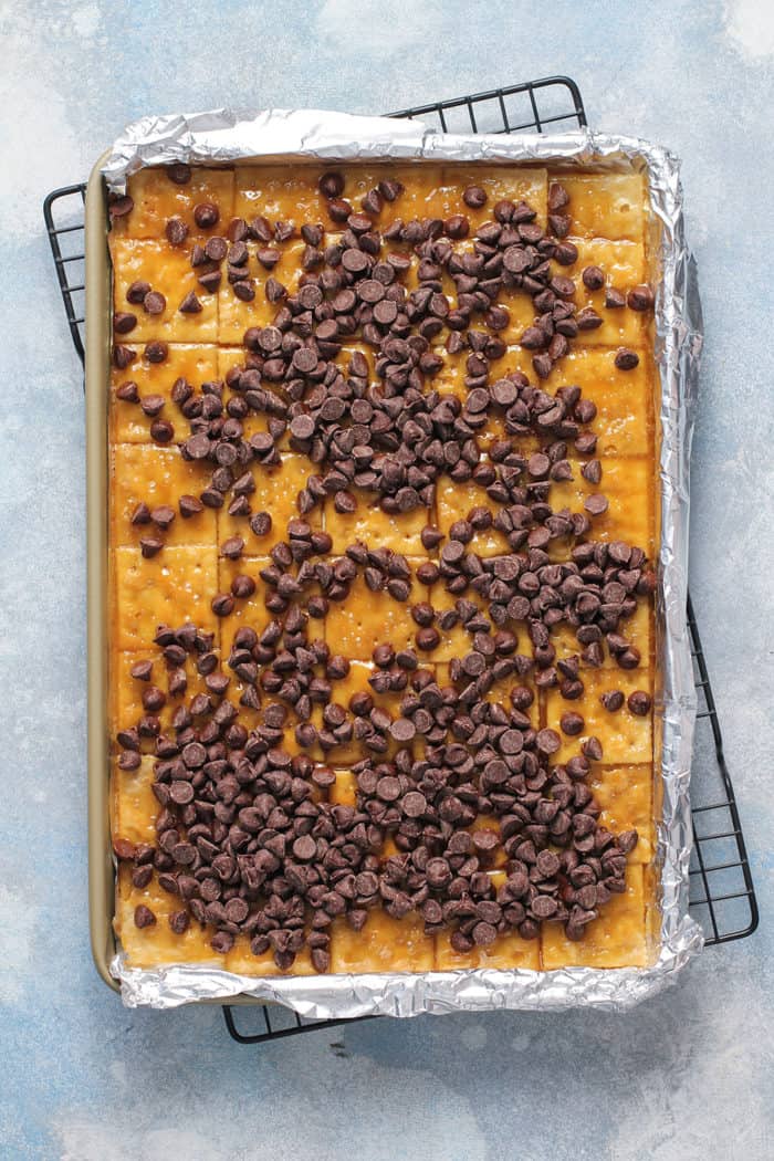 Chocolate chips sprinkled on top of saltine toffee in a foil-lined sheet pan