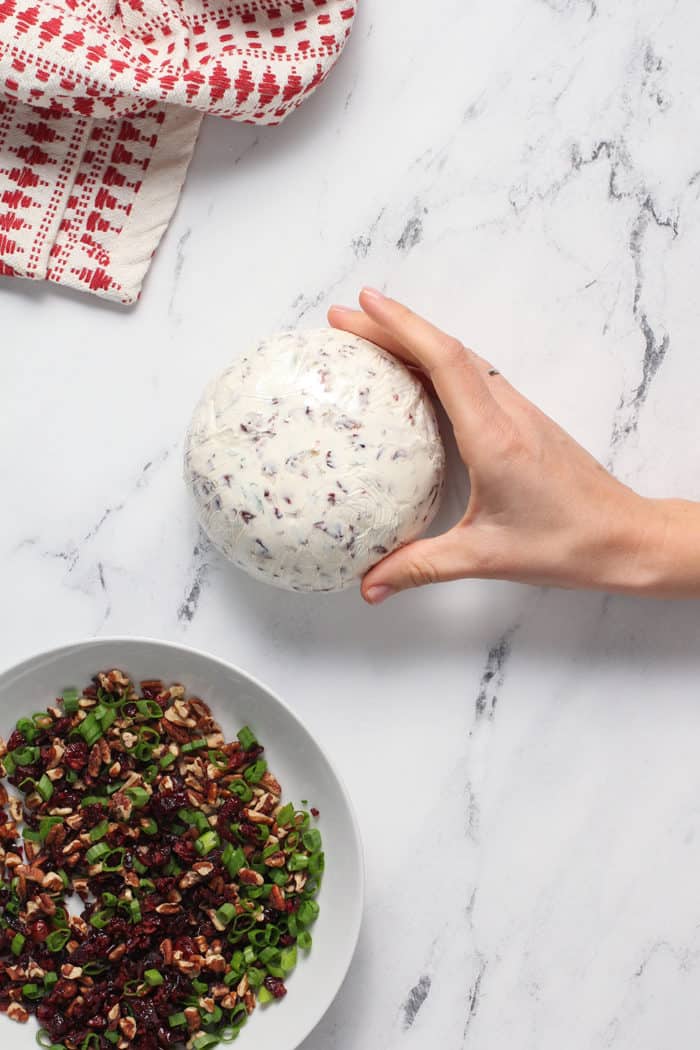 Formed cranberry goat cheese ball next to a plate with chopped pecans, cranberries, and green onions for coating the outside