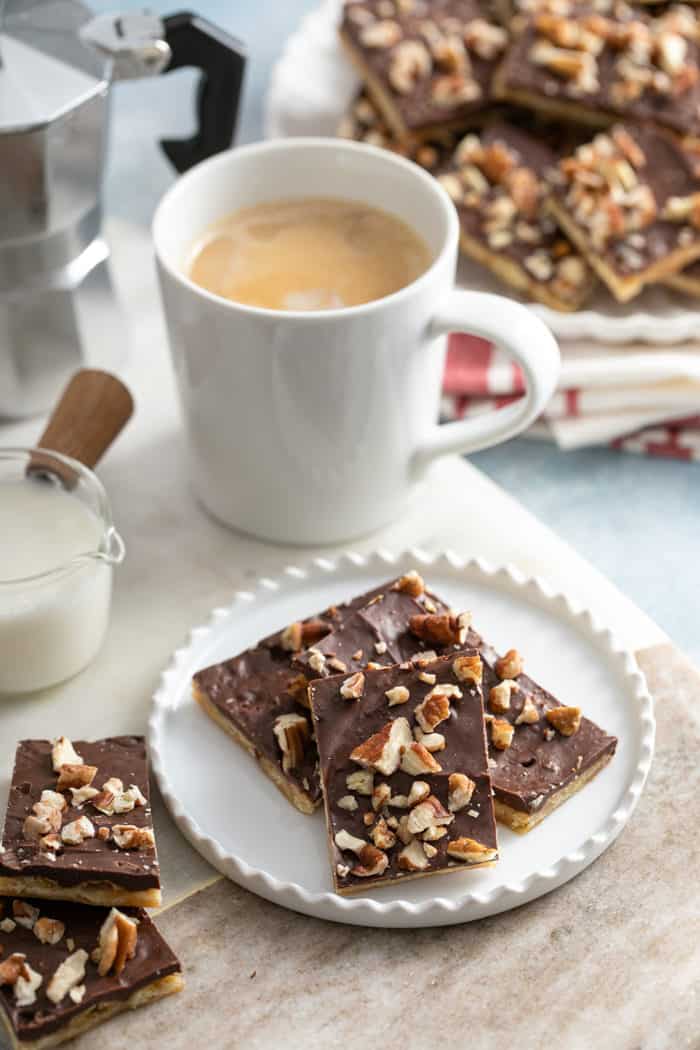 Two pieces of saltine toffee arranged on a white plate next to a cup of coffee