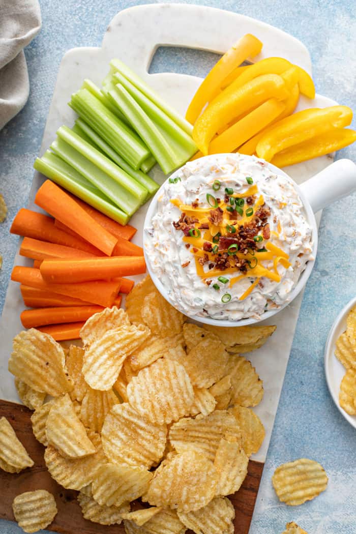 Overhead view of a white bowl filled with loaded baked potato dip on a platter next to potato chips and fresh veggies