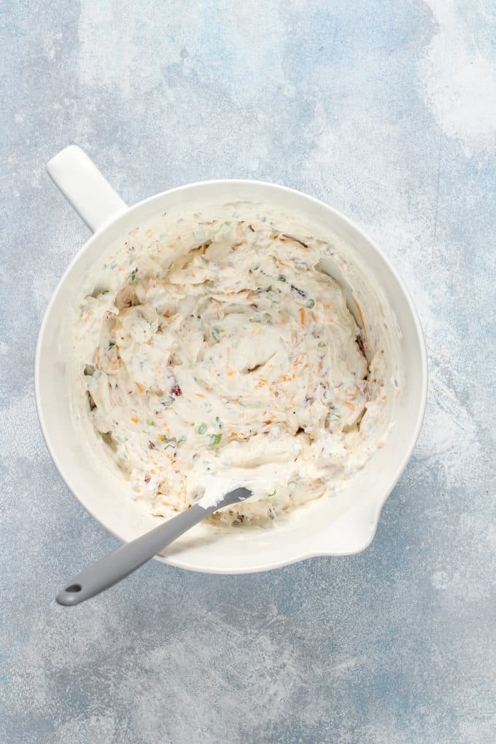 Spatula stirring together loaded baked potato dip in a white mixing bowl