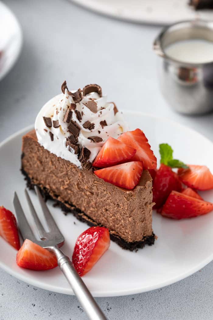 Slice of chocolate cheesecake with a bite taken out of it on a white plate, garnished with whipped cream and strawberries