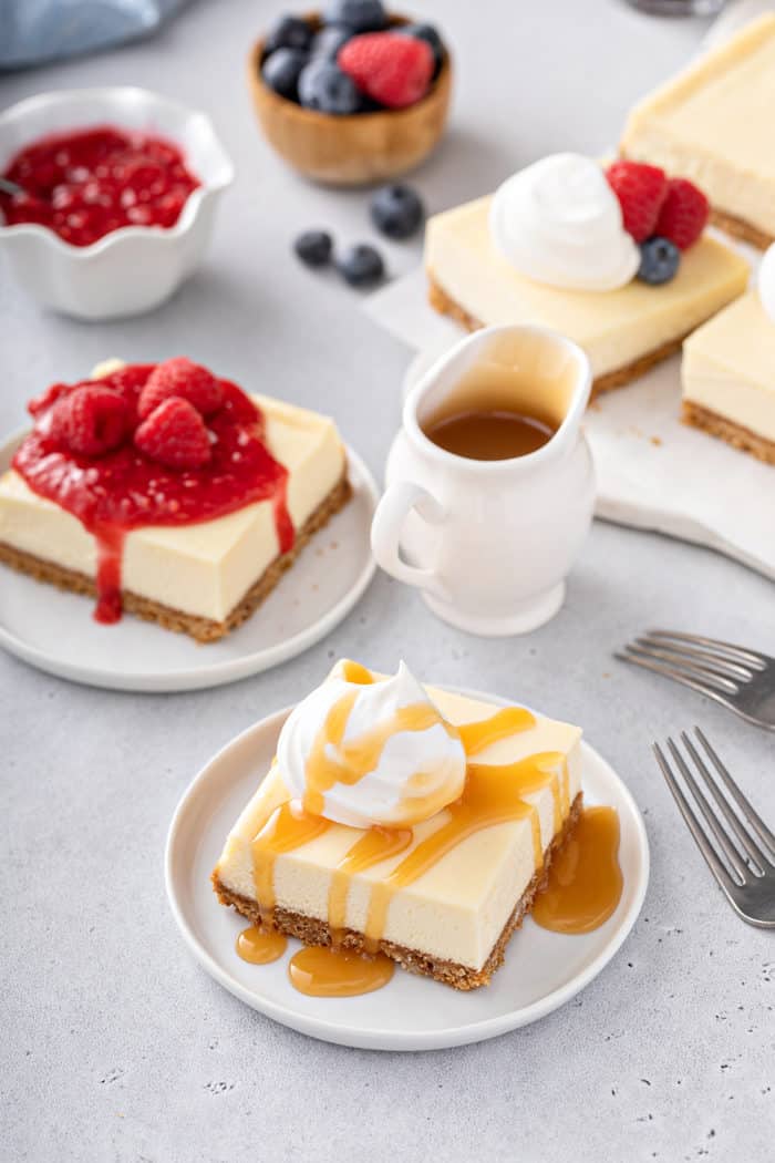 Plated cheesecake bars topped with various toppings: caramel, raspberry sauce, and fresh fruit