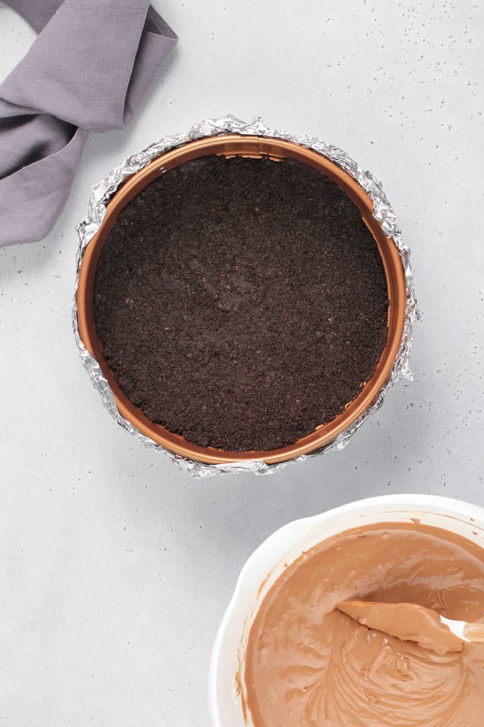 Oreo cookie crust in a springform pan next to a bowl of chocolate cheesecake filling