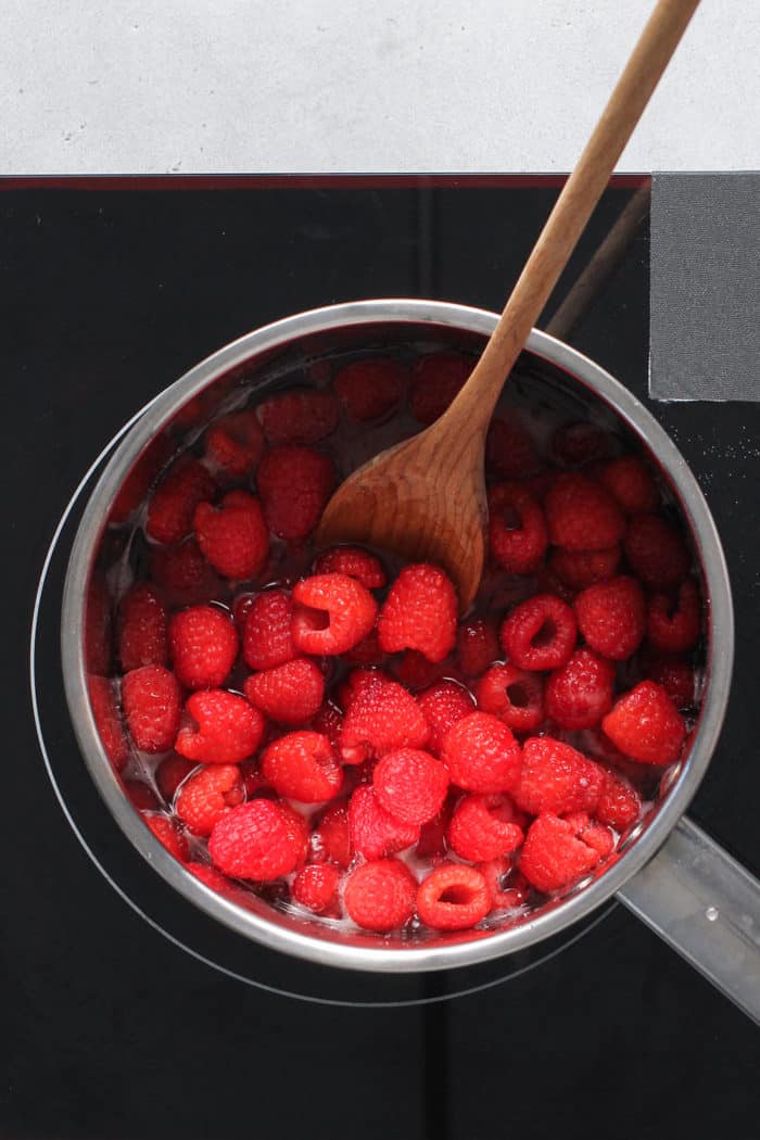 Raspberries being stirred by a wooden spoon in a metal saucepan