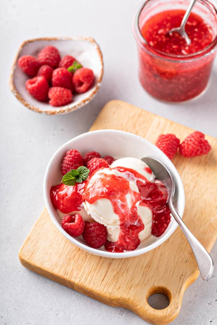 White bowl filled with vanilla ice cream topped with raspberry sauce. Fresh raspberries and a jar of raspberry sauce are in the background