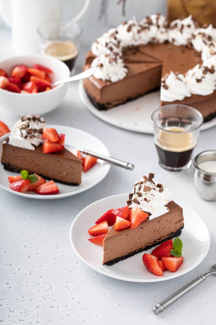 Two plated slices of chocolate cheesecake, garnished with whipped cream and strawberries, with a sliced cheesecake in the background