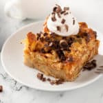 Slice of chocolate chip bread pudding on a white plate, topped with whipped cream and chocolate shavings