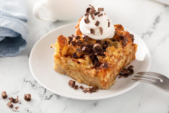 Slice of chocolate chip bread pudding on a white plate, topped with whipped cream and chocolate shavings