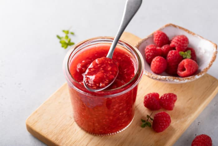 Jar of raspberry sauce and fresh raspberries set on a wooden cutting board