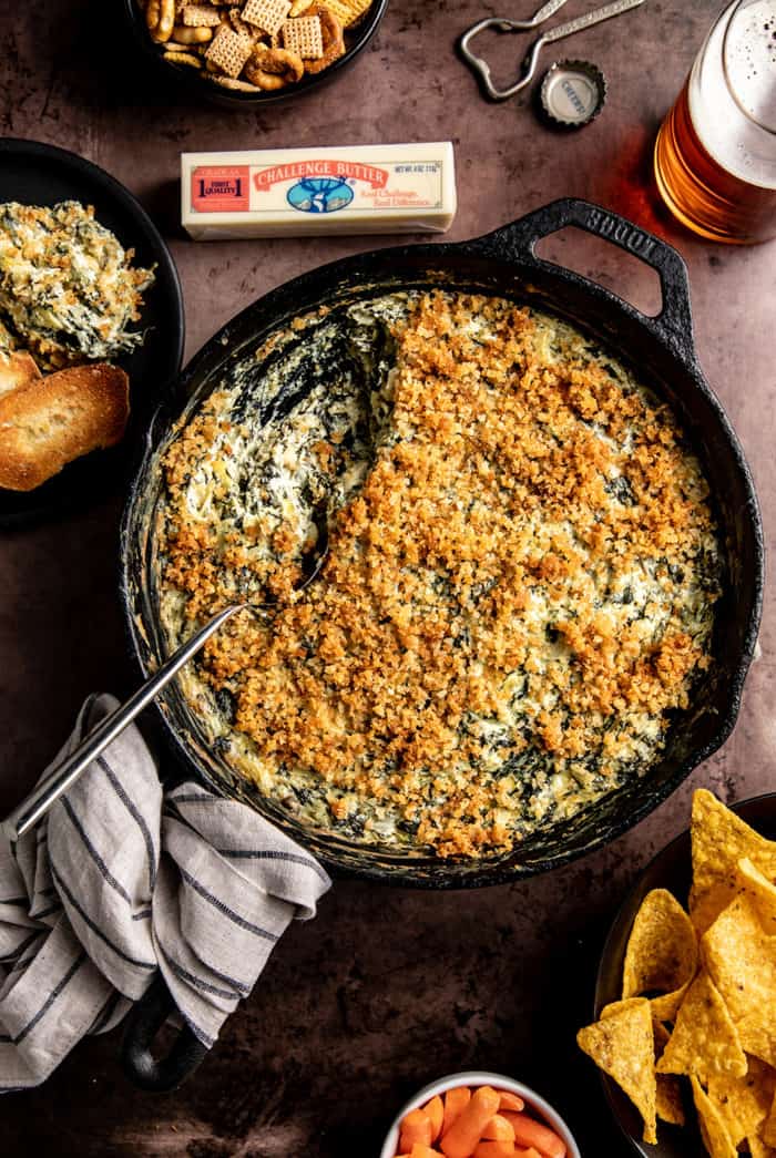 Skillet of spinach artichoke dip surrounded by beer and chips. Some of the dip has been served and a spoon is resting in the skillet